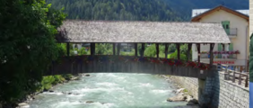 Bild einer alten Brücke mit Ziegeldach. Im Hintergrund ein Gebirgsbach und der Fuß eines Berges, der mit Nadelbäumen bewaldet ist.