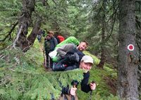 Mehrere Wanderer mit Rucksäcken und Trekkingstöcken auf einem schmalen Waldpfad, rechts ein Baum mit rotem und weißem Wanderzeichen.