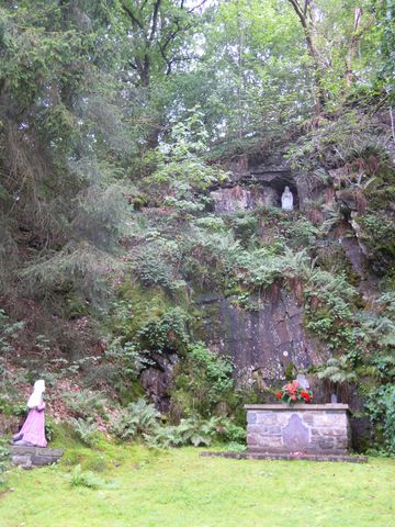 Eine steinerne Statue einer Frau in rosa Kleid steht links im Bild, während rechts eine Felswand mit einer Statue der Jungfrau Maria und üppiger Vegetation zu sehen ist. Im Vordergrund befindet sich ein kleiner Altar mit Blumen.