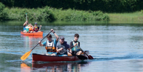 Zwei Kanus mit jeweils mehreren Personen, die auf einem ruhigen Gewässer paddeln, umgeben von grüner Ufervegetation