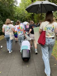 Mehrere Personen gehen auf einem Weg, eine trägt einen Regenschirm, andere T-Shirts mit Aufdrucken wie 'BORN THIS WAY' und 'ALLE SIND WILLKOMMEN'