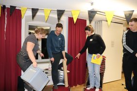 Das Bild zeigt junge Menschen, die auf Stühlen stehen und Gegenstände in der Hand balancieren. Sie versuchen eine schwierige Aufgabe zu lösen, ohne den Boden zu berühren.