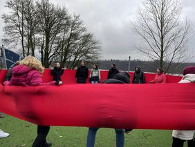 Mehrere Personen stehen im Kreis auf einem Spielfeld und halten gemeinsam ein großes, rotes Tuch gespannt.