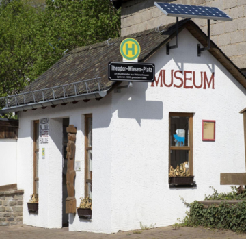 Eingang zu einem Museum mit rotem Schriftzug "MUSEUM". Das Gebäude hat ein schräges Dach und eine Solaranlage. Neben dem Eingang befindet sich ein Schild mit dem Namen "Theodor-Wiesen-Platz".