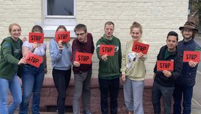 Gruppe von acht Jugendlichen steht vor einer Hauswand und hält jeweils ein rotes Schild mit der Aufschrift 'STOP'.