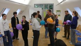 Mehrere Jugendliche stehen in einem Raum mit Holzboden und weißen Wänden, einige halten bunte Luftballons, im Hintergrund sind Stühle und Fenster zu sehen.