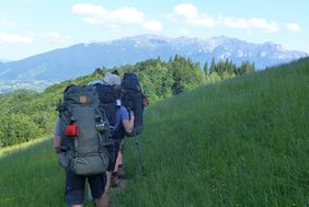 Wanderer mit großen Rucksäcken gehen auf einem grasbewachsenen Hang, im Hintergrund bewaldete Hügel und Berge unter blauem Himmel.