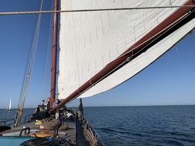 Blick vom Deck eines Segelschiffs mit gehisster Segel und mehreren Personen, die auf das offene Meer blicken.