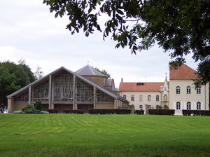 Moderne Kirche mit Glasfront und angrenzendem historischen Gebäude in einer grünen Parklandschaft.