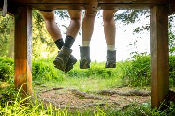 Zwei Paar Füße in Wanderschuhen hängen von einer Holzbank, umgeben von grüner Wiese und Bäumen.