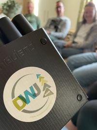 Nahaufnahme eines schwarzen Ordners mit einem runden Aufkleber der DWJ NRW, im Hintergrund unscharfe Personen in einem Sitzkreis