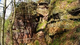 Ds Bild zeigt eine steile Felsenwand, die schön vom Sonnenlicht beleuchtet wird. Es ist ein sonniger Herbsttag und man läuft einen schmalen Pfad entlang der Felsen. Links geht es weit herunter, rechts geht es steil nach oben.