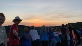 Das Bild zeigt junge Menschen, die gegen eine Mauer lehnen und den Sonnenuntergang anschauen. Die Landschaft ist ein Mischwald und ein Tal im rot-orangenen Licht der untergegangenen Sonne.