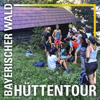 Ein Bild von einer Gruppe junger Menschen, die sich vor einer alten Hütte sammelt. Sie haben kurze Sachen an, weil es heiß ist. Die Hütte ist im Schatten der umliegenden Nadelbäume verborgen.