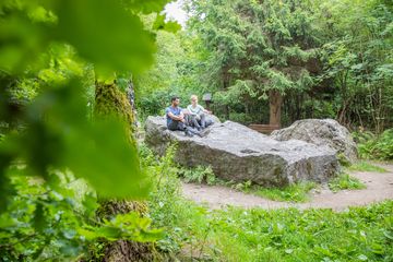 Zwei Personen sitzen auf einem großen Felsen in einem Waldgebiet, umgeben von grünen Pflanzen und Bäumen.