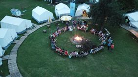 Luftaufnahme eines Zeltlagers mit mehreren weißen Zelten und einer großen Gruppe von Menschen, die im Kreis um ein Lagerfeuer sitzen.
