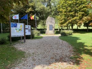 Ein Park mit einem Weg, der zu einem großen Gedenkstein führt. Links stehen Informationsschilder und Flaggen von Europa, Belgien und anderen Ländern. Umgeben von Bäumen und Grünflächen.
