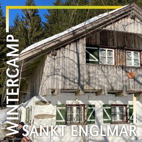 Bild einer Alpenhütte im Sonnenlicht erhellt. Es liegt schnee und hinter der Hütte stehen große Nadelbäume. Der Himmel ist wunderbar blau. Die Fensterläden der Hütte sind offen und grün.