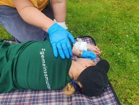 Person liegt auf einer karierten Decke auf dem Gras, eine andere Person mit blauen Handschuhen hält die Hand der Liegenden