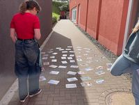 Zwei Personen stehen auf einem gepflasterten Weg zwischen zwei Gebäuden, auf dem zahlreiche Karten oder Bilder verteilt liegen.