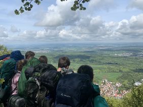 Das Bild zeigt eine Gruppe Jugendlicher, die einen atemberaubenden Blick hinunter auf die Schwäbische Landschaft genießen. Sie hocken auf einem Felsen am Albtrauff der Schwäbischen Alb. Sie haben große Wanderrucksäcke. Es ist sonnig und man kann viele Kilometer weit blicken.