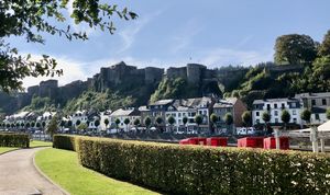 Blick auf eine historische Burganlage über einem Fluss, umgeben von grünen Hügeln und einem malerischen Ufer mit Häusern und Bäumen. Im Vordergrund eine gerade geschnittene Hecke.
