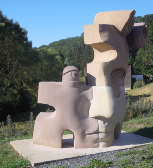 Skulptur in abstrakter Form, die Gesichtsmerkmale darstellt, aus hellem und dunklem Material, im Freien auf einer Wiese platziert.