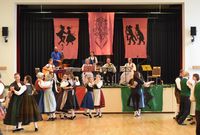 Mehrere Paare in traditioneller Tracht tanzen in einem Saal vor einer Bühne, auf der Musiker mit verschiedenen Instrumenten spielen, hinter ihnen hängen drei große Banner mit tanzenden Figuren und einem Herzmotiv.
