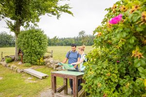 Zwei Personen, ein Mann und eine Frau, stehen an einem Tisch im Freien und betrachten eine Karte. Um sie herum ist eine grüne Landschaft mit Bäumen und Blumen.