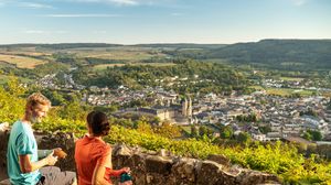 Paar sitzt auf einer Aussichtskanzel, blickt auf eine malerische Stadtlandschaft umgeben von Hügeln und Wäldern. Die Sonne scheint, der Himmel ist klar.