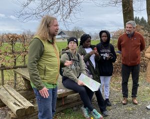 Gruppe von fünf Personen bei einer Outdoor-Aktivität, eine Person spricht, während andere zuhören. Eine Person hält eine Karte, die Umgebung ist ländlich.