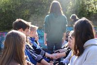 Jugendliche sitzen im Freien in zwei Reihen einander gegenüber, eine Person steht zwischen ihnen mit dem Rücken zur Kamera.