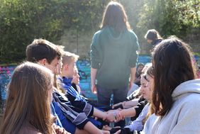 Jugendliche sitzen im Freien in zwei Reihen einander gegenüber, eine Person steht zwischen ihnen mit dem Rücken zur Kamera.