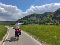 Zwei Radfahrer auf einem asphaltierten Donaurad-Weg neben einer Wiese mit gelben Blumen, im Hintergrund bewaldete Felsen unter leicht bewölktem Himmel. Die Kalkfelsen des Donautales beugen sich mächtig über dem Tal auf.
