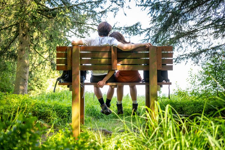 Paar sitzt auf einer Holzbank im Grünen, umgeben von Bäumen und Gras. Die Person links hat einen Arm um die Schulter der Person rechts gelegt.