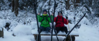 Zwei Personen in Winterkleidung sitzen auf einer Holzbank im Schnee, vor ihnen stehen mehrere Skier im Schnee.