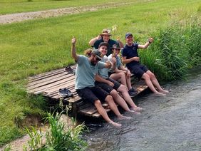Fünf Personen sitzen auf einem Holzsteg am Wasser, haben die Füße im Wasser und zeigen Daumen nach oben